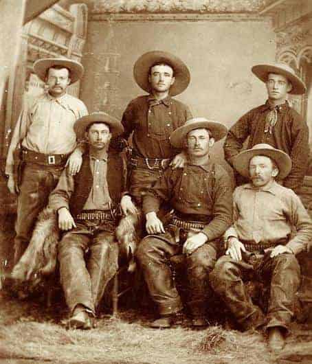 Six Texas Rangers dressed in 1860s cowboy clothes including hats, rifles, and bullet belts, pose for a portrait